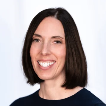 A woman with short hair in a blue shirt, standing against a neutral background with a friendly expression.