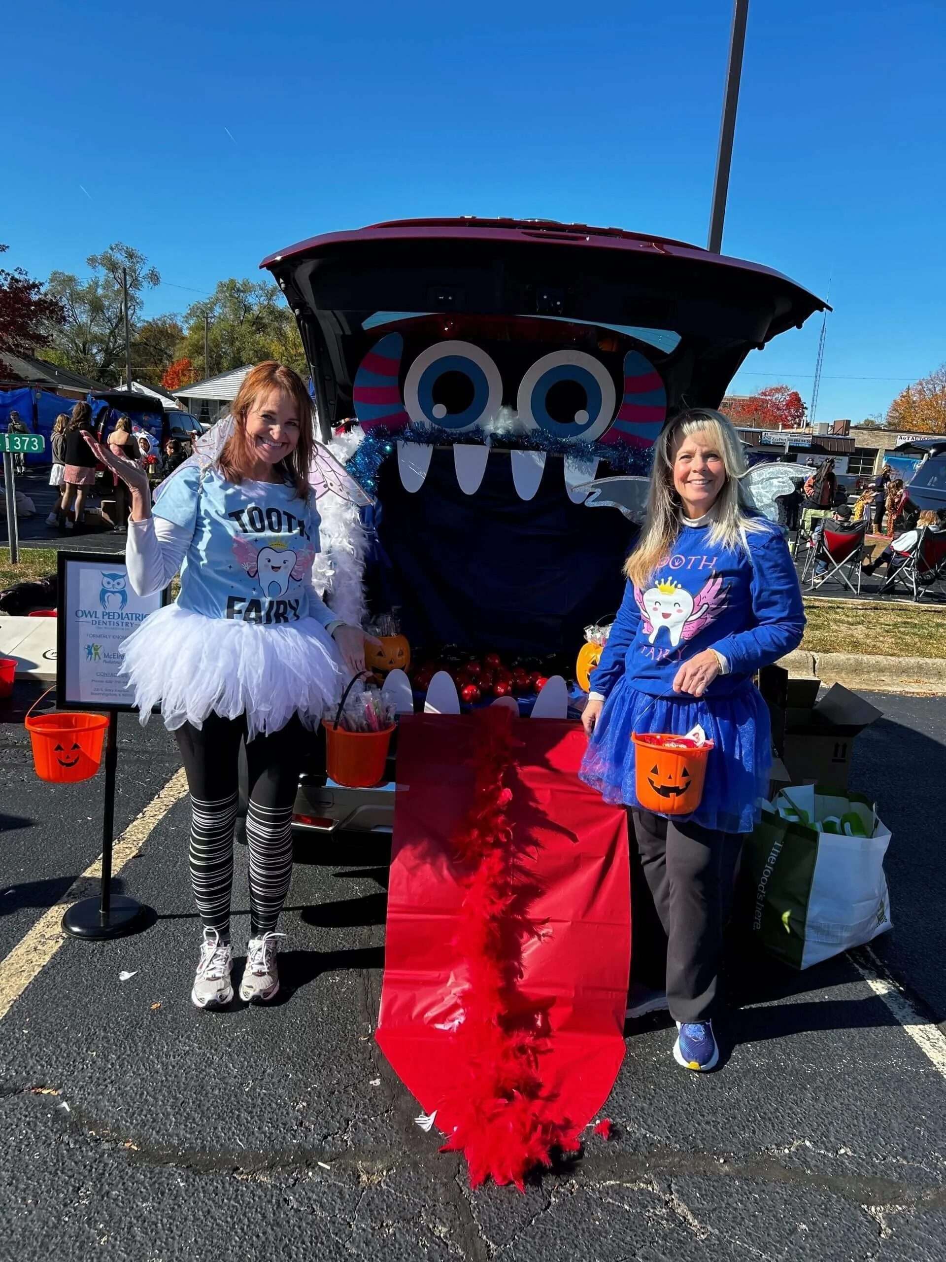 Two women dressed as tooth fairies stand in front of a car decorated with large eyes and teeth for Halloween. They hold pumpkin buckets, and the car trunk is filled with treats and a red cloth resembling a tongue.